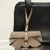 Estuche para lentes de ecocuero color beige con textura granulada, diseño de borde festoneado y cierre con broches metálicos dorados. Incluye una correa corta con mosquetón para colgar en bolsos o carteras.