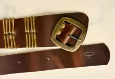 Faja de cuero marrón con hebilla metálica cuadrada en tono bronce envejecido y detalles decorativos de varillas metálicas verticales. Presenta un diseño artesanal con logo grabado en el extremo.