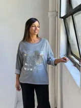Remera gris de manga larga con cuello redondo y diseño holgado. Presenta un aplique frontal rectangular con detalles de lentejuelas plateadas y estampado tipográfico.