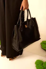Cartera tipo tote de cuero sintético color negro con textura granulada. Tiene doble asa corta, cierre con broche metálico dorado y costuras en contraste color blanco.