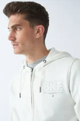 Campera de felpa color blanco hueso, con capucha y cordón ajustable, cierre frontal con cremallera, bolsillos laterales, logo y arte estampado en el pecho.