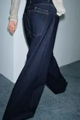 Jeans de tiro alto con pierna ancha, confeccionados en denim azul oscuro. Presentan cinco bolsillos, trabillas en la cintura y un cinturón ajustable del mismo tejido con hebilla metálica. Incluyen pespuntes marcados a tono y cierre frontal con cremallera y botón.