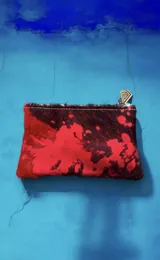 Monedero de cuero vacuno con pelo, de diseño rectangular y cierre superior, con estampa abstracta en tonos rojos y marrones. Cuenta con un dije metálico con forma geométrica en el cierre.