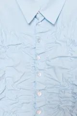Camisa de algodón con cuello solapa y manga larga. Presenta un diseño con frunces elásticos en el panel frontal y bajo asimétrico. Cierre frontal con botones.