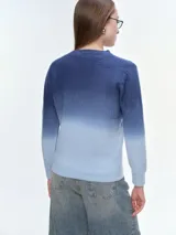 Buzo tejido de punto con diseño degradé que transiciona de azul oscuro a celeste claro. Presenta cuello redondo, mangas largas y puños acanalados, con un corte holgado y relajado.
