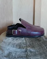 Zuecos de cuero vegano en color bordó con acabado texturizado. Presentan un diseño de punta cerrada con una tira ajustable con hebilla metálica que puede usarse en el tobillo o sobre el empeine.