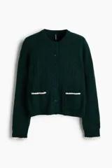 Saco corto de punto trenzado fino en color verde oscuro, con cuello redondo, cierre frontal de botones y bolsillos delanteros. Presenta detalles de ribetes festoneados en color crema en los bolsillos y en los puños.