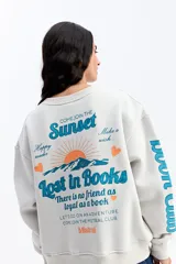 Buzo de algodón color marrón chocolate, con cuello redondo y diseño holgado. Presenta una estampa gráfica en la espalda con motivos de atardecer y montañas, junto con texto decorativo. Además, incluye una estampa en la manga con letras grandes.