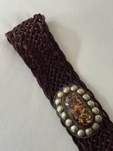 Cinto trenzado de cuero color chocolate con hebilla ovalada de metal envejecido, decorada con una piedra central jaspeada y tachas metálicas alrededor.