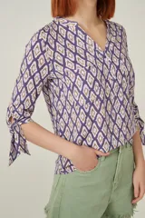 Blusa de viscosa con estampado geométrico en tonos violeta y beige. Tiene cuello mao, botones frontales y mangas 3/4 con lazo en los puños.