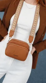 Bandolera de tamaño compacto confeccionada en cuero sintético color marrón claro. Presenta dos compartimentos principales con cierre y una correa ancha ajustable con diseño geométrico estampado.
