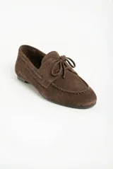 Mocasín plano de gamuza color marrón oscuro, estilo náutico o driving shoe, con costura visible en la puntera y lazo decorativo en el empeine.