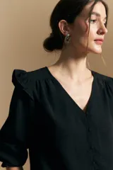 Camisa negra de manga larga con escote en V y cierre frontal de botones. Presenta detalles de volados en los hombros, frunces en la espalda y puños elastizados que le otorgan una silueta relajada y fluida.