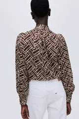 Blusa de manga larga en tejido transparente con estampado geométrico en tonos marrón y blanco. Presenta cuello elevado con frunces y puños ajustados con abertura y botón.