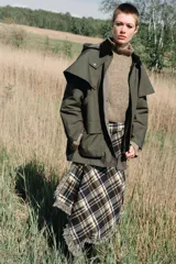 Trench de estilo encerado color verde militar con cuello de solapa en pana marrón, capucha extraíble y capa superior. Presenta cierre frontal con cremallera y botones a presión ocultos, bolsillos delanteros de plastrón con solapa y mangas largas con trabillas.