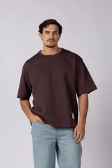 Remera de corte oversize color marrón oscuro, con cuello redondo y mangas cortas amplias. Presenta una etiqueta pequeña en el ruedo inferior.