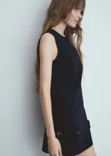 Vestido corto sin mangas de corte recto, con cuello redondo y cierre de cremallera invisible en la parte posterior. Presenta detalles de botones decorativos en el bajo y aberturas laterales.