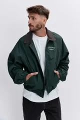 Cazadora de estilo urbano en color verde oscuro, con cuello de pana en tono marrón, cierre frontal con cremallera y puños elásticos. Presenta un diseño holgado con bordado pequeño en el pecho.