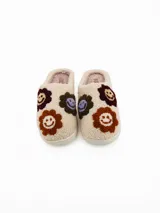 Pantuflas de estar por casa tipo mule, confeccionadas en tejido de peluche color crema. Presentan un diseño con apliques bordados de caritas sonrientes en tonos marrones, bordó y lila. Cuentan con una suela plana de goma y forro interior suave.