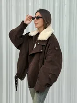 Campera de gamuza color marrón con cuello de corderito sintético en color crema. Presenta un diseño oversize con cierre frontal, solapas con broches metálicos y una tira ajustable en el ruedo.