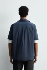 Camisa de manga corta con cuello bowling, confeccionada en popelín de algodón. Presenta un corte relajado, cierre frontal con botones y aberturas laterales en el bajo.
