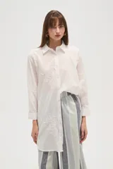 Camisa blanca de manga larga, corte holgado y largo, con cuello camisero y botones frontales. Presenta bordados florales en tono blanco sobre el tejido.