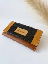 Billetera de cuero con diseño tricolor en tonos negro y marrón. Presenta una solapa central con textura acolchada y un parche con el logo de la marca, flanqueada por paneles laterales con textura de reptil. Incluye cierre con broche y monedero externo.