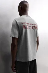 Camiseta relaxed fit de algodón, color gris melange con mangas en un tono de gris más claro. Tiene cuello redondo, manga corta, bordado frontal y texto estampado en la espalda a contraste. Incluye un parche con logo en el bajo.