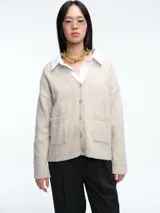 Cardigan de punto color beige con cierre frontal de botones, cuello en V y dos bolsillos delanteros. Presenta mangas largas con puños acanalados y un diseño de corte holgado.