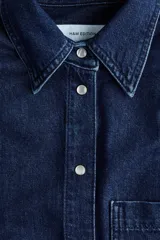 Camisa en denim de algodón con hombreras, cuello clásico, botones a presión nacarados, bolsillo frontal aplicado y bajo redondeado.