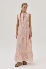 Vestido largo de algodón color rosa con estampado floral en tonos beige y dorado. Tiene cuello alto con lazo ajustable, corte imperio, falda escalonada con volados y diseño sin mangas.