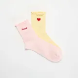 Pack de dos pares de medias de algodón acanalado, una en color rosado con el texto 'Amour' bordado y otra en color amarillo con un corazón rojo bordado. Ambas cuentan con detalles de volantes en los bordes superiores.