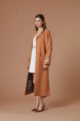 Saco largo color tostado con diseño de corte recto y silueta relajada. Presenta cierre frontal con botones, bolsillos laterales amplios y un parche decorativo en la manga izquierda. El interior cuenta con un forro estampado artístico.