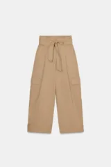 Pantalón de tiro alto estilo paperbag en color beige, con pinzas frontales y cinturón de la misma tela. Presenta bolsillos laterales tipo cargo con solapa y pernera ajustable con botones en el bajo.