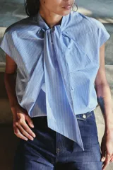 Camisa sin mangas confeccionada en algodón, con cuello redondo y detalle de lazada frontal. Presenta un diseño de corte holgado con bajo redondeado y cierre posterior mediante abertura y botones.