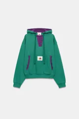 Sudadera con capucha Fruit of the Loom en colaboración con Zara, de color verde con detalles en morado en el interior de la capucha, la cartera de botones y los bordes del bolsillo frontal tipo canguro. Presenta mangas largas, cordones ajustables y un parche con el logo en el centro.