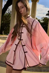 Camisa de algodón de estilo oriental, color rosado con ribetes marrón oscuro, cuello mao, mangas largas amplias con puños y cierre frontal con nudos chinos.