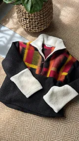 Buzo de corderito combinado de The Mood, con cuello alto, cierre frontal parcial de cremallera y diseño de bloques de color. Presenta estampado a cuadros en tonos rojo, amarillo y negro en la parte superior y mangas, y cuerpo negro liso con puños blancos de corderito.