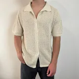 Camisa de tejido de punto calado en color beige, con cuello polo, cierre frontal de botones y mangas cortas.