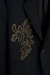 Blazer de corte relajado confeccionada en tejido con lana, color negro. Presenta cuello con solapas de muesca, cierre frontal de un botón y bolsillos de plastrón en la cadera. Destaca por su bordado en hilo dorado con motivos vegetales en la parte frontal.
