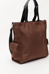 Bolso tipo tote color negro, con doble asa superior y correa desmontable. Presenta un bolsillo frontal con solapa, bolsillos laterales y un detalle colgante decorativo con tiras de colores.