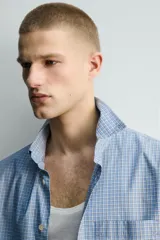 Camisa de hombre de manga larga, corte relajado (relaxed fit), color celeste con patrón de cuadros pequeños en azul y rojo. Presenta cuello abotonado (button-down), bolsillo de parche en el pecho y cierre frontal de botones.
