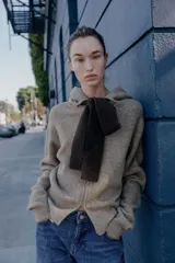 Chaqueta de punto con capucha, manga larga y cierre frontal con cremallera. Presenta un diseño en color beige con un lazo decorativo a contraste en color marrón oscuro en la zona del cuello.