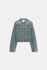Chaqueta denim entallada de cuello redondo y manga larga, en tono azul desgastado. Presenta un diseño inspirado en estilos militares con múltiples filas de botones metálicos plateados en el frente y cierre con ganchos ocultos.
