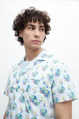 Camisa de manga corta con estampado de flores y palmeras en tonos azules, verdes y naranjas sobre fondo blanco. Tiene cuello americano, bolsillo en el pecho y botones personalizados.
