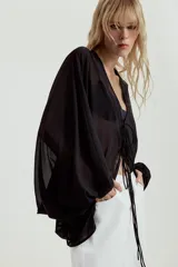 Top de crepe blanco con estampado de lunares negros, escote en V y cierre frontal con finas lazadas. Presenta mangas largas abullonadas con puños elásticos y bajo redondeado.
