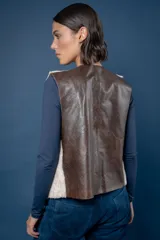 Chaleco de piel sintética en color beige, con cierre frontal mediante hebillas metálicas y tiras de cuero marrón que cuelgan como detalle decorativo.