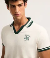 Remera polo comfort de algodón color marfil, con cuello en V verde oscuro con rayas blancas, mangas cortas con puños rayados y estampa de escudo bordada en el pecho.