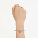 Brazalete rígido de la colección Idyllia, con diseño de media caña adornado con cristales transparentes. Presenta un dije central en forma de corazón tipo medallón, con baño en tono oro y una zirconia rosa en una cara, y pavé de cristales con baño de rodio en la otra.