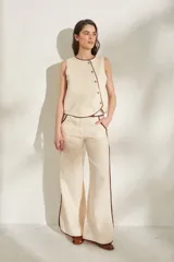 Pantalon de lino color beige claro con detalles de ribetes en tono marron oscuro en los bolsillos, trabillas y laterales de las piernas. Presenta corte recto y amplio con tiro medio.